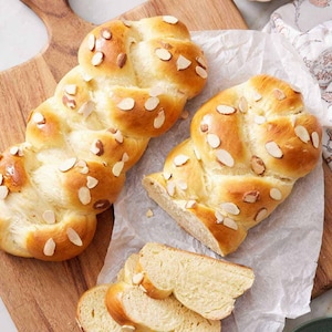 May include: Braided bread topped with sliced almonds, displayed on a wooden cutting board with slices in front. A small bowl of red dyed eggs is in the background, suggesting a festive occasion.