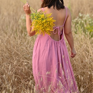 May include: A pink, open-back dress with spaghetti straps. The dress is worn in a field of tall, dry grass. A bouquet of yellow flowers is held in the person's hand. The dress has a flowing, relaxed fit, suitable for warm weather.