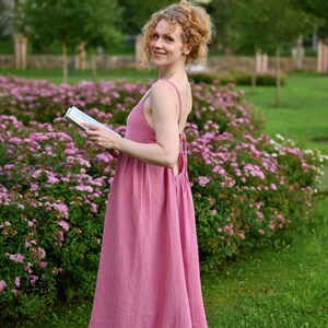 Puede incluir: Una mujer con un vestido largo rosa con tirantes finos, en un jard&iacute;n con flores rosas. El vestido es de tela fluida y texturizada. Sostiene un libro y sonr&iacute;e. El vestido es de color rosa empolvado.