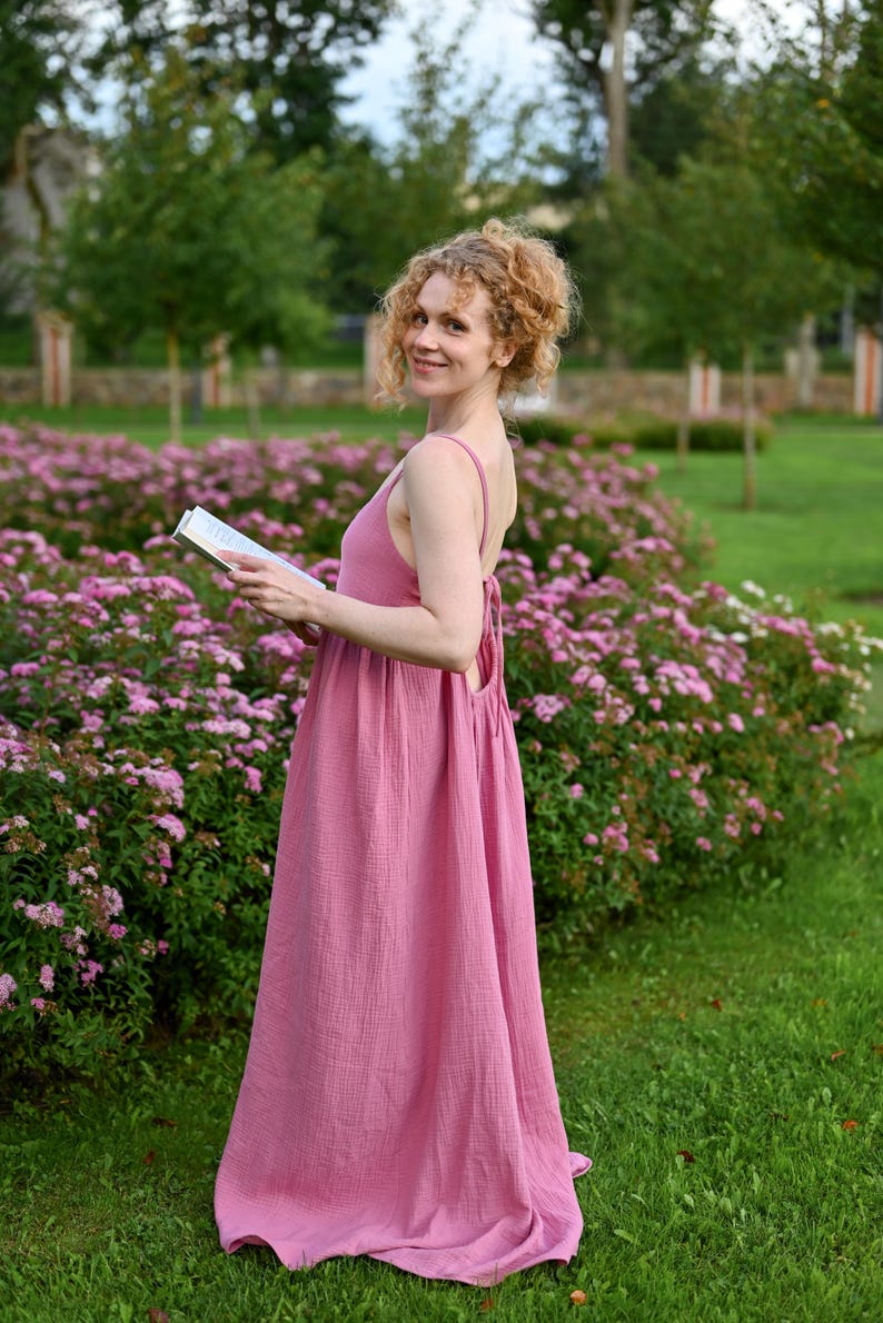 Puede incluir: Una mujer con un vestido largo rosa con tirantes finos, en un jard&iacute;n con flores rosas. El vestido es de tela fluida y texturizada. Sostiene un libro y sonr&iacute;e. El vestido es de color rosa empolvado.