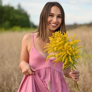 Puede incluir: Una persona con un vestido rosa sostiene un ramo de flores amarillas en un campo. El vestido tiene cuello en V y tirantes finos. El fondo es un campo de trigo y un cielo nublado.
