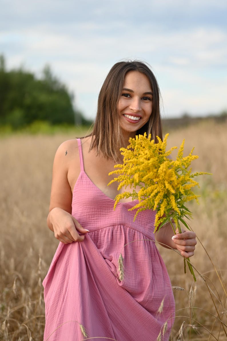 Puede incluir: Una persona con un vestido rosa sostiene un ramo de flores amarillas en un campo. El vestido tiene cuello en V y tirantes finos. El fondo es un campo de trigo y un cielo nublado.