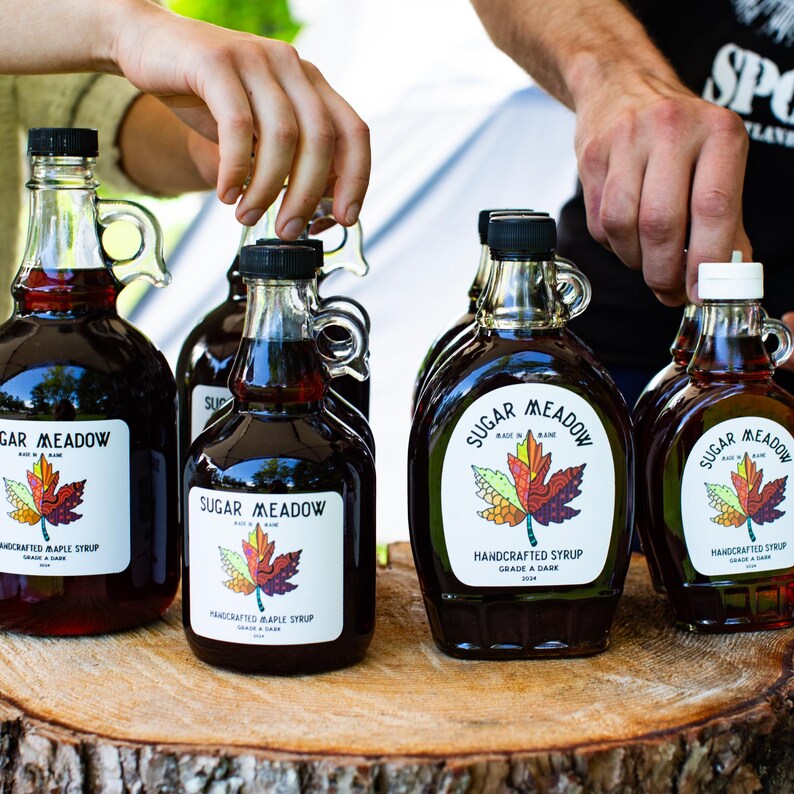 May include: Several glass bottles of maple syrup with black caps and handles are arranged on a wooden surface. Each bottle has a label that reads "Sugar Meadow Handcrafted Maple Syrup" with a colorful leaf design. The syrup is a dark amber color.