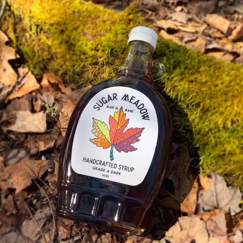 May include: A clear glass bottle of Sugar Meadow handcrafted syrup, Grade A Dark, with a colorful maple leaf design on the label. The bottle is filled with dark syrup and sits on a bed of leaves and moss.