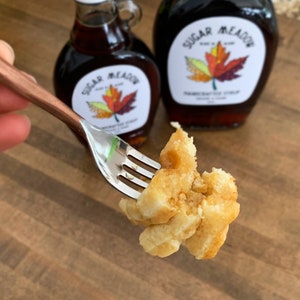 K&ouml;nnte beinhalten: Eine Gabel h&auml;lt ein St&uuml;ck goldbraunen Waffel mit etwas Ahornsirup darauf. Im Hintergrund stehen zwei Flaschen Ahornsirup, beide mit dem Etikett "Sugar Meadow" und einem Ahornblatt-Design.