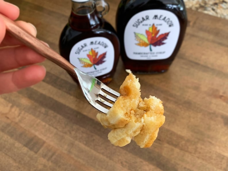 K&ouml;nnte beinhalten: Eine Gabel h&auml;lt ein St&uuml;ck goldbraunen Waffel mit etwas Ahornsirup darauf. Im Hintergrund stehen zwei Flaschen Ahornsirup, beide mit dem Etikett "Sugar Meadow" und einem Ahornblatt-Design.