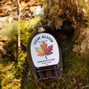 May include: A dark glass bottle of maple syrup with a white label featuring a colorful maple leaf design. The label reads "Sugar Meadow Handcrafted Syrup Grade A Dark 2025" and "Made in Maine". The bottle has a white cap and a handle.