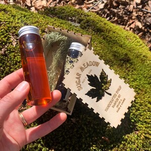 May include: A sampler set of handcrafted syrup in small glass bottles with silver lids. The bottles are displayed in a wooden box with the words "Sugar Meadow" and "Handcrafted Syrup" laser-cut into the wood. The syrup is Grade A Dark & Amber.