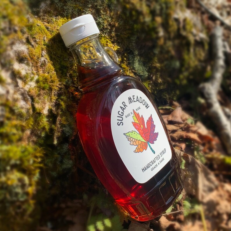 K&ouml;nnte beinhalten: Eine Glasflasche Ahornsirup mit einem wei&szlig;en Etikett, auf dem "Sugar Meadow Made in Maine Handcrafted Syrup Grade A Dark" steht. Das Etikett zeigt eine farbenfrohe Illustration eines Ahornblattes.