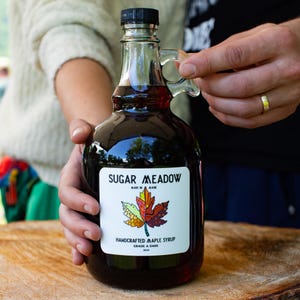 May include: A glass bottle of dark maple syrup with a label that reads "Sugar Meadow, Made in Maine, Handcrafted Maple Syrup, Grade A Dark, 2024". The label features a colorful maple leaf design.