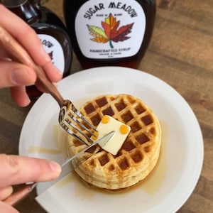 May include: A golden-brown waffle on a white plate with a pat of butter being cut with a fork and knife. Maple syrup is drizzled around the waffle. Two bottles of maple syrup are in the background, one with a label that reads "Sugar Meadow Handcrafted Syrup".