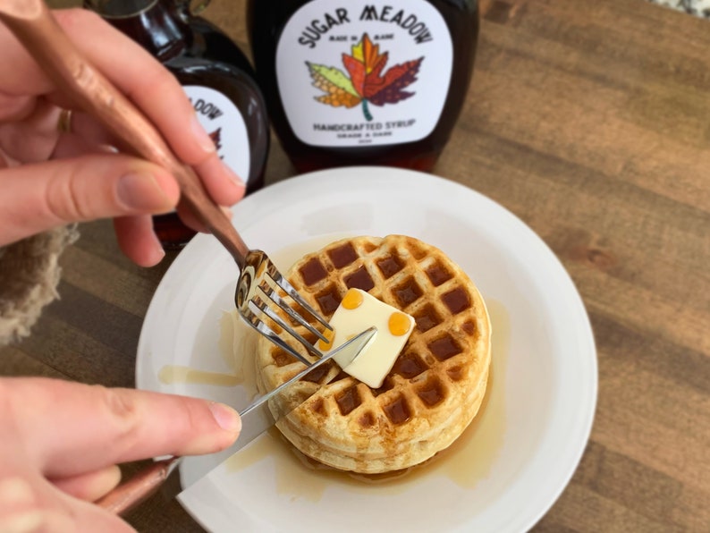 May include: A golden-brown waffle on a white plate with a pat of butter being cut with a fork and knife. Maple syrup is drizzled around the waffle. Two bottles of maple syrup are in the background, one with a label that reads "Sugar Meadow Handcrafted Syrup".