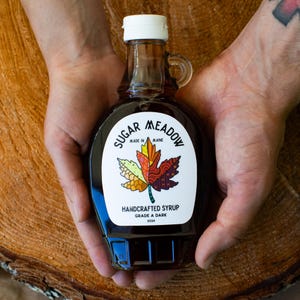 K&ouml;nnte beinhalten: Eine Glasflasche mit dunklem Ahornsirup mit einem wei&szlig;en Etikett, auf dem "Sugar Meadow Made in Maine" steht und ein farbenfrohes Ahornblatt-Design abgebildet ist. Das Etikett tr&auml;gt au&szlig;erdem die Aufschrift "Handgemachter Sirup G&uuml;teklasse A Dunkel 2024".