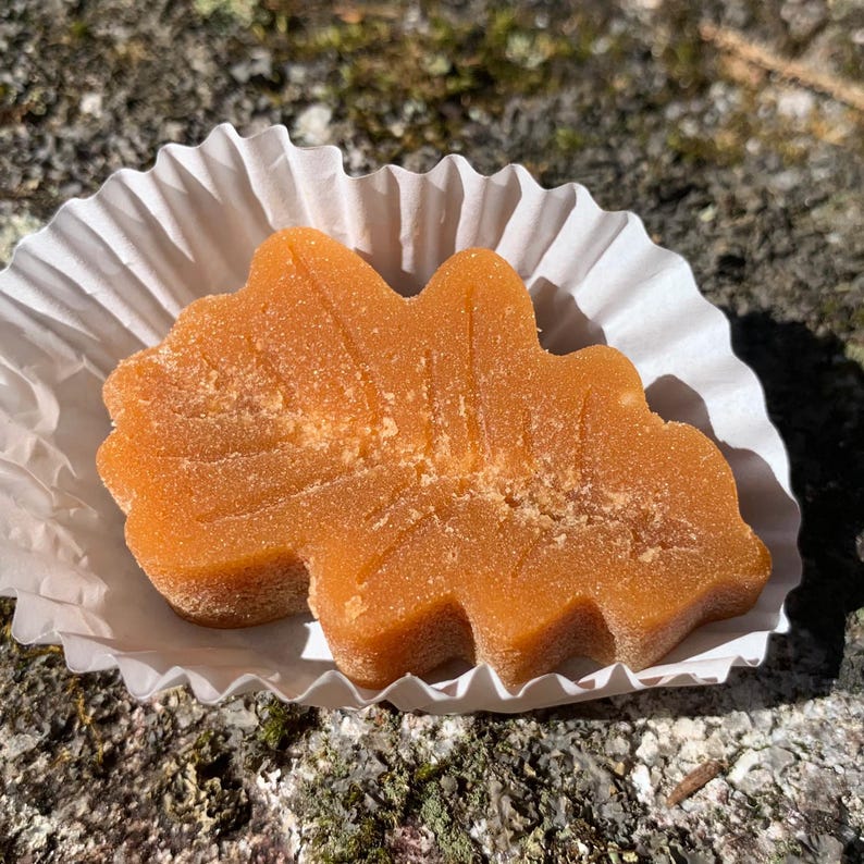May include: A maple leaf-shaped candy made of maple syrup, in a white paper cup. The candy is a light brown color and has a slightly grainy texture.
