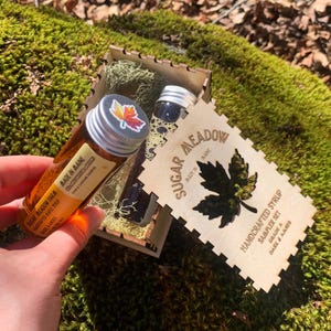 May include: A maple syrup sampler set in a wooden box. The box is laser-cut with the words "Sugar Meadow" and a maple leaf design. Two small glass bottles of syrup are visible, one with a label that reads "Made in Maine."