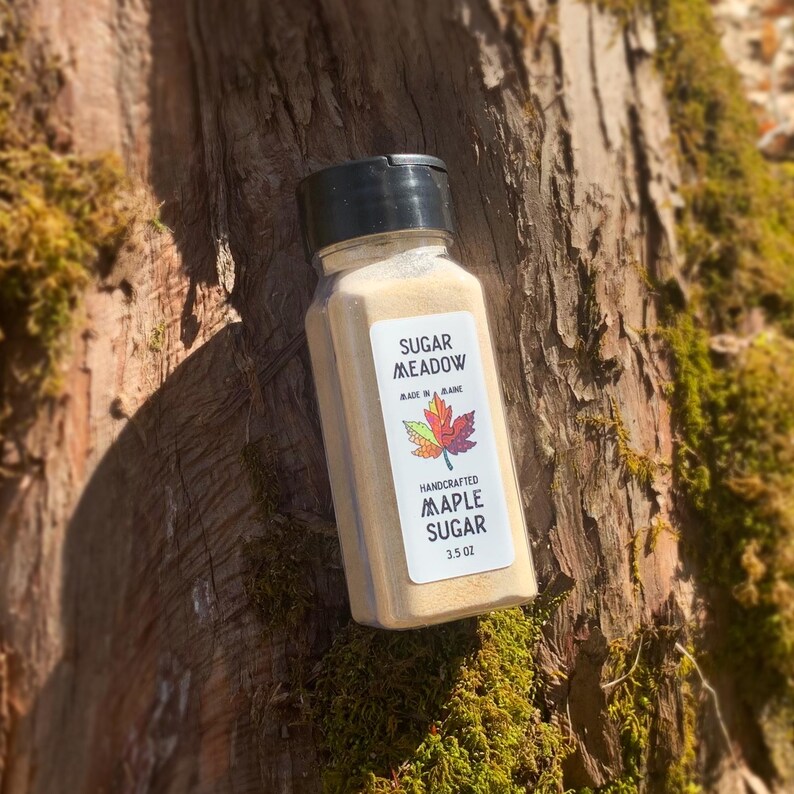 May include: A small glass jar with a brown lid filled with a light brown powder. The jar has a white label with the text "Sugar Meadow" and "Handcrafted Maple Sugar 3.5 oz" printed on it.