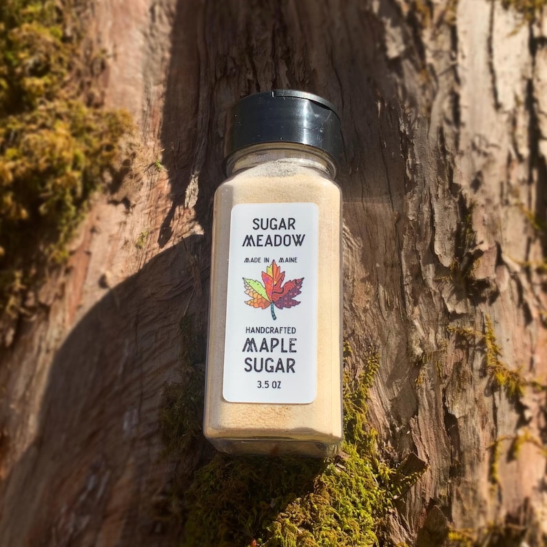 May include: A glass jar of maple sugar with a white label that says "Sugar Meadow, Made in Maine, Handcrafted Maple Sugar, 3.5 oz". The label also features a colorful maple leaf.