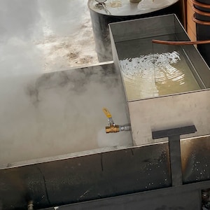 May include: A stainless steel evaporator with a yellow valve and copper tubing. Steam rises from the liquid inside the evaporator. A metal barrel is visible in the background. The image depicts a maple syrup production setup.