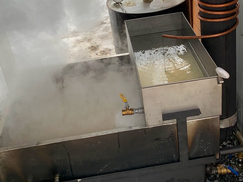May include: A stainless steel evaporator with a yellow valve and copper tubing. Steam rises from the liquid inside the evaporator. A metal barrel is visible in the background. The image depicts a maple syrup production setup.
