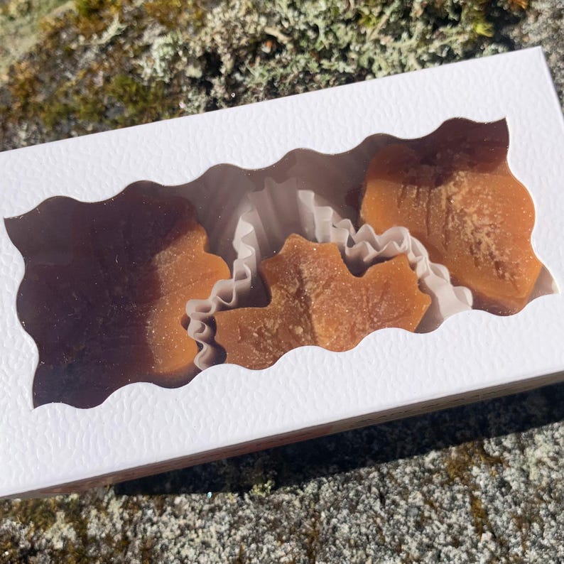 May include: A white box with a clear window showing three maple leaf shaped candies in individual white paper cups. The candies are a light brown color and have a slightly grainy texture.