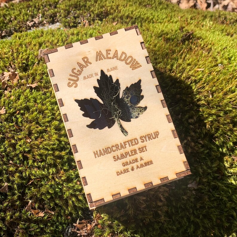 May include: A wooden box with the text "Sugar Meadow Made in Maine" and "Handcrafted Syrup Sampler Set". The box features a dark maple leaf design. The grade is listed as Dark & Amber.