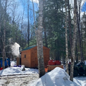 May include: A wooden sugar shack with a green roof and a chimney emitting steam, set in a snowy forest. Red containers sit on a wooden pallet, and a four-wheeler is parked nearby. A blue tent is also visible.