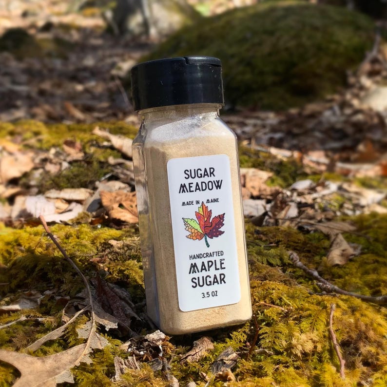 May include: A clear glass jar with a black lid filled with light brown maple sugar. The jar has a white label with the text "Sugar Meadow" in black lettering, "Made in Maine" in smaller black lettering, a colorful maple leaf illustration, and "Handcrafted Maple Sugar 3.5 oz" in black lettering.
