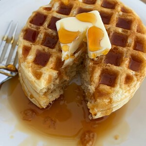 May include: A golden-brown waffle, cut to reveal its fluffy interior, topped with a pat of butter and drizzled with syrup. The waffle sits on a white plate, with a fork to the side. A breakfast treat.