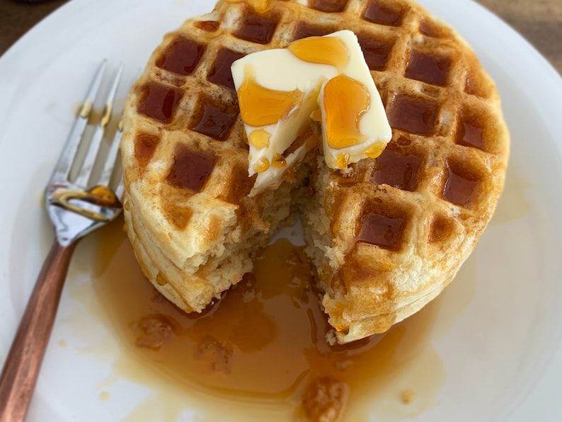 May include: A golden-brown waffle, cut to reveal its fluffy interior, topped with a pat of butter and drizzled with syrup. The waffle sits on a white plate, with a fork to the side. A breakfast treat.