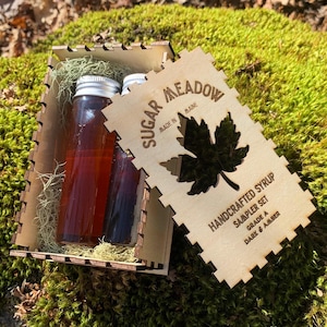May include: A wooden box set containing two glass bottles of syrup. The bottles are filled with a dark amber liquid and have silver lids. A wooden tag with the text "Sugar Meadow" and "Handcrafted Syrup" is included.