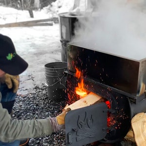 May include: A person adding firewood to a stainless steel evaporator, with flames visible. Steam rises from the evaporator, and sparks fly. The scene suggests maple syrup production, with a black cap and gloves.