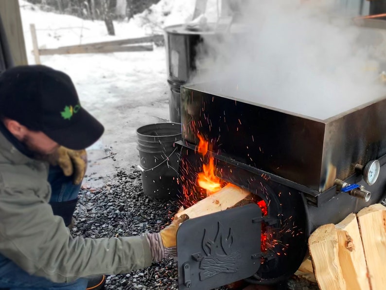 May include: A person adding firewood to a stainless steel evaporator, with flames visible. Steam rises from the evaporator, and sparks fly. The scene suggests maple syrup production, with a black cap and gloves.