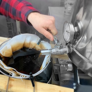 May include: A person in a red and black plaid shirt is adjusting a valve on a stainless steel brewing tank. A mesh bag filled with a dark liquid is suspended in a container. A gauge displays pressure readings.