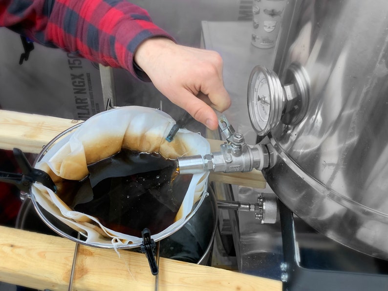 May include: A person in a red and black plaid shirt is adjusting a valve on a stainless steel brewing tank. A mesh bag filled with a dark liquid is suspended in a container. A gauge displays pressure readings.