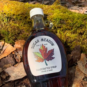 K&ouml;nnte beinhalten: Eine Glasflasche mit dunklem Ahornsirup mit einem Etikett, auf dem "Sugar Meadow Made in Maine Handcrafted Syrup Grade A Dark" steht. Das Etikett zeigt ein farbenfrohes Ahornblatt-Design.