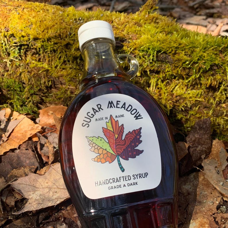 K&ouml;nnte beinhalten: Eine Glasflasche mit dunklem Ahornsirup mit einem Etikett, auf dem "Sugar Meadow Made in Maine Handcrafted Syrup Grade A Dark" steht. Das Etikett zeigt ein farbenfrohes Ahornblatt-Design.