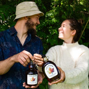 May include: Two glass bottles of maple syrup with the label "Sugar Meadow" and a maple leaf design. The bottles are held by two people in front of a green leafy background. The syrup is a rich amber color.