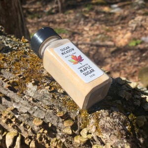 May include: A small glass jar filled with light brown maple sugar. The jar has a white label with the text "Sugar Meadow" and "Handcrafted Maple Sugar 3.5 oz".