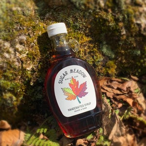 K&ouml;nnte beinhalten: Eine Glasflasche mit dunklem Ahornsirup mit einem wei&szlig;en Etikett, auf dem "Sugar Meadow" und "Handgemachter Sirup G&uuml;teklasse A dunkel" steht. Das Etikett zeigt auch ein farbenfrohes Ahornblatt.