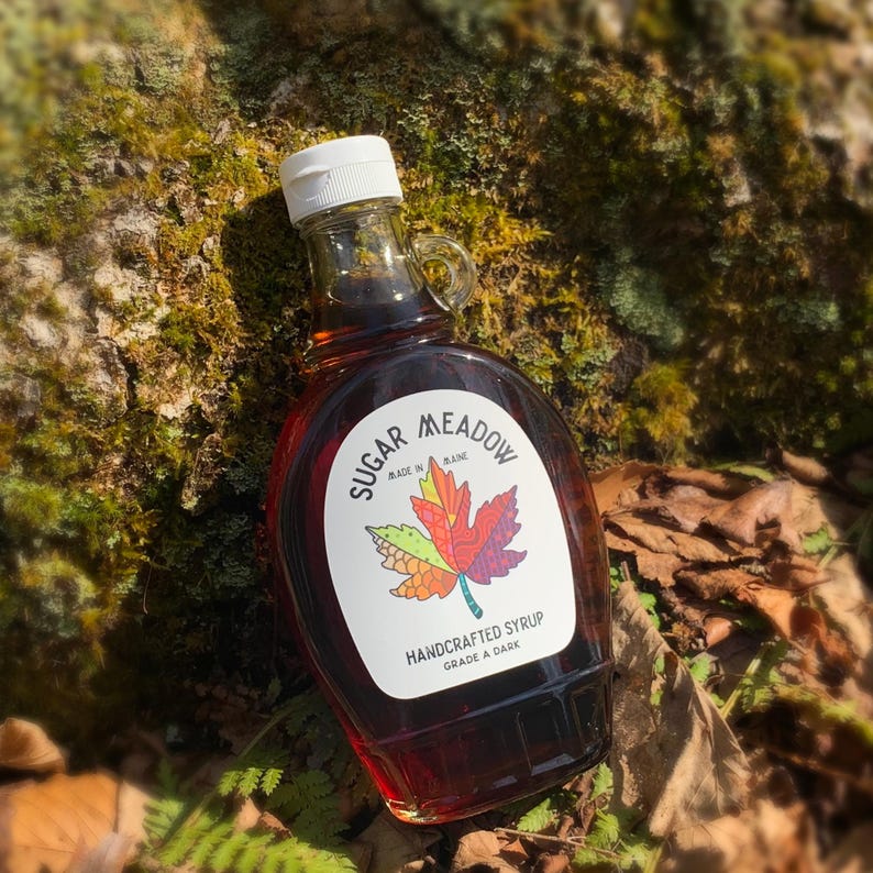 K&ouml;nnte beinhalten: Eine Glasflasche mit dunklem Ahornsirup mit einem wei&szlig;en Etikett, auf dem "Sugar Meadow" und "Handgemachter Sirup G&uuml;teklasse A dunkel" steht. Das Etikett zeigt auch ein farbenfrohes Ahornblatt.