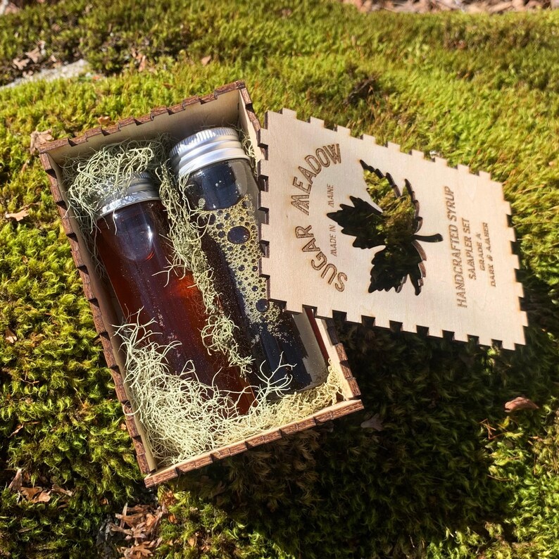 May include: A wooden box containing two small glass bottles of syrup with silver lids, nestled in dried moss. The box has a laser-cut lid with the words "Sugar Meadow" and "Handcrafted Syrup Sampler Set". The syrup is dark amber in color.