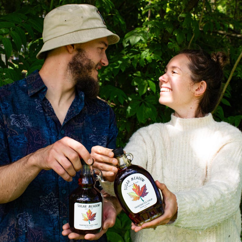 May include: Two glass bottles of maple syrup with the label "Sugar Meadow" and a maple leaf design. The bottles are held by two people in front of a green leafy background. The syrup appears dark amber in color.