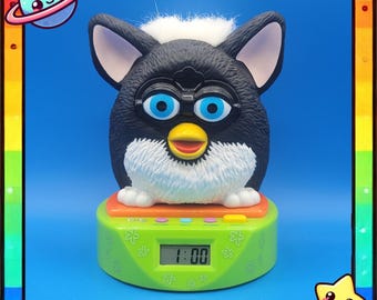 Puede incluir: Un despertador estilo Furby en blanco y negro con ojos azules y un pico amarillo. El reloj está sobre una base verde con una pantalla digital que muestra la hora 1:00. El reloj tiene un mechón de pelo blanco en la parte superior y está sobre un fondo azul.