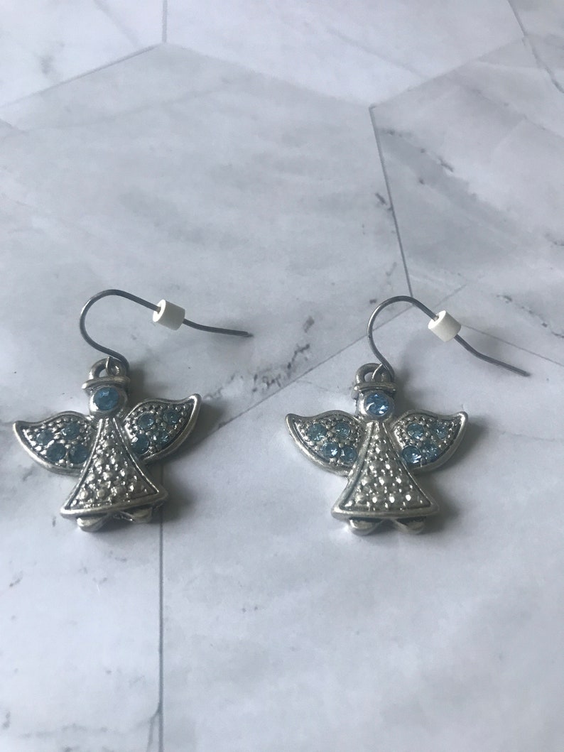 May include: A pair of silver angel earrings with blue crystal accents. The earrings have a hook closure.