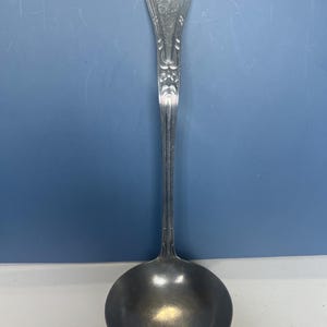 Puede incluir: Un cucharón plateado con un cuenco redondo y un mango largo y ornamentado. El mango presenta un diseño floral decorativo en la parte superior y a lo largo de su longitud. El cucharón está diseñado para servir sopas y guisos.
