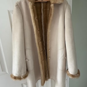 May include: A cream-coloured coat with a faux fur collar and trim. The coat has a button-down front and two pockets. The interior lining is a light brown colour. The coat is hanging on a wooden hanger.