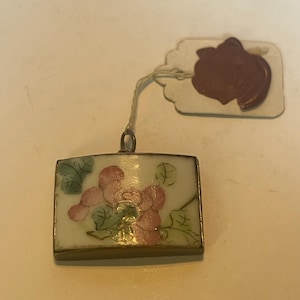Może przedstawiać: Mały, kwadratowy wisiorek z kwiatowym wzorem. Wisiorek wykonany jest z białej porcelany z malowanymi różowymi i zielonymi kwiatami. Wisiorek jest przymocowany do złotego łańcuszka.