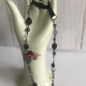 Puede incluir: Un collar negro y plateado con cuentas negras, cuentas transparentes y eslabones de cadena plateados. El collar se muestra en una mano de maniquí blanca.