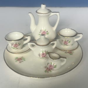 May include: A miniature porcelain tea set with a teapot, creamer, sugar bowl, two teacups, and saucers. The set is white with gold trim and delicate pink rose floral designs. The tea set is arranged on a matching white tray.
