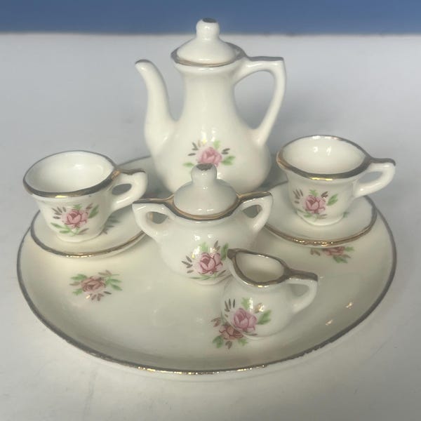 May include: A miniature porcelain tea set with a teapot, creamer, sugar bowl, two teacups, and saucers. The set is white with gold trim and delicate pink rose floral designs. The tea set is arranged on a matching white tray.